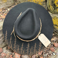 A hand-burned black wool hat resting on weathered timber, its brim etched with a dense, silhouetted forest.
