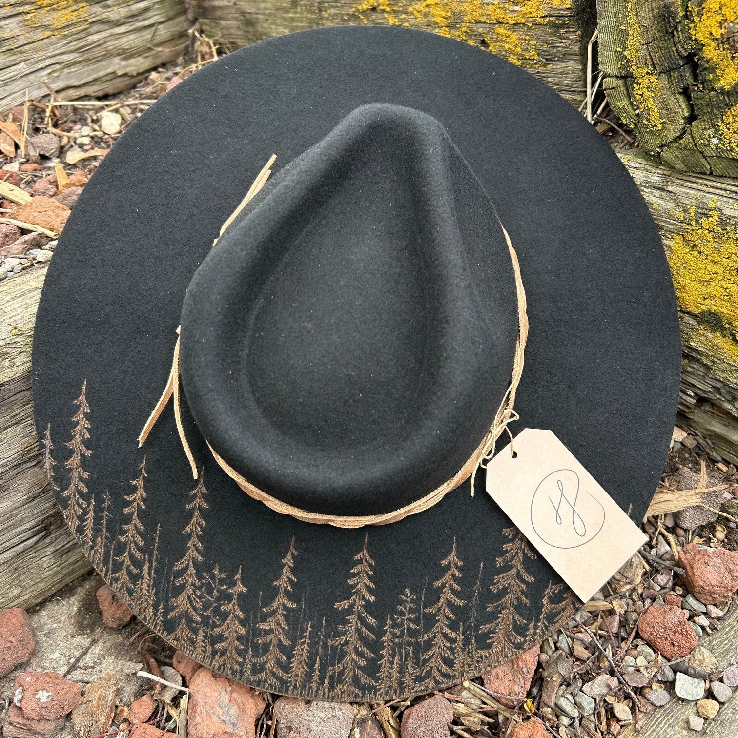 A hand-burned black wool hat resting on weathered timber, its brim etched with a dense, silhouetted forest.