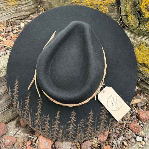 A hand-burned black wool hat resting on weathered timber, its brim etched with a dense, silhouetted forest.