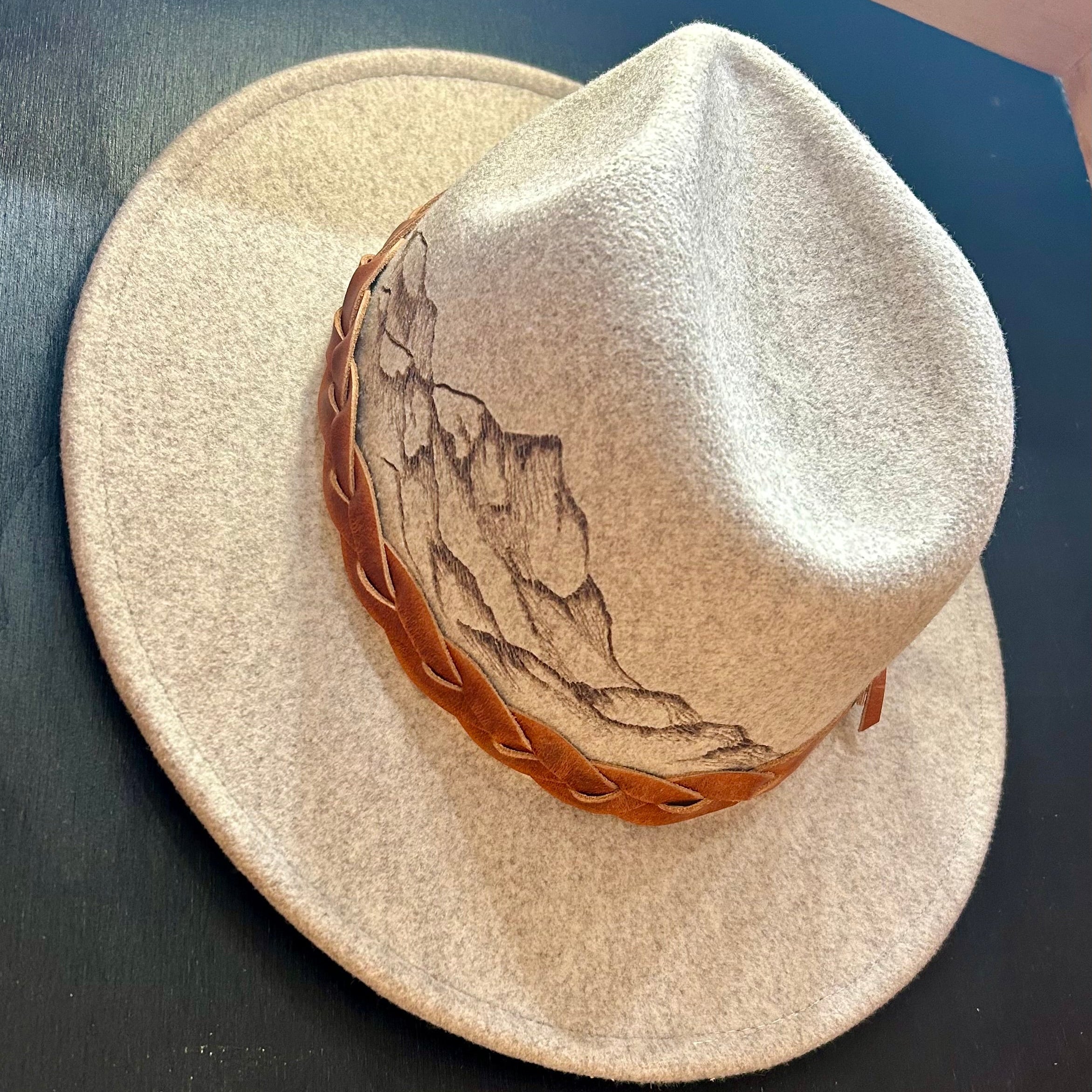 Hand-burned mountain landscape etching along the crown of a custom felt hat. A jagged, minimalist peak silhouette designed for high-altitude style.