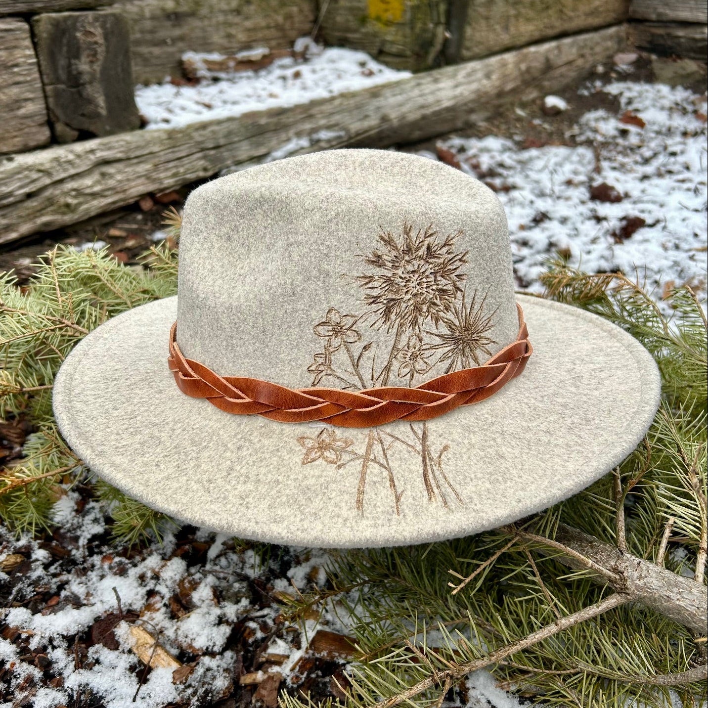 A tangle of wild, hand-etched floral stems erupts from the brim to the crown of a light grey felt hat, accompanied by a handmade leather band.